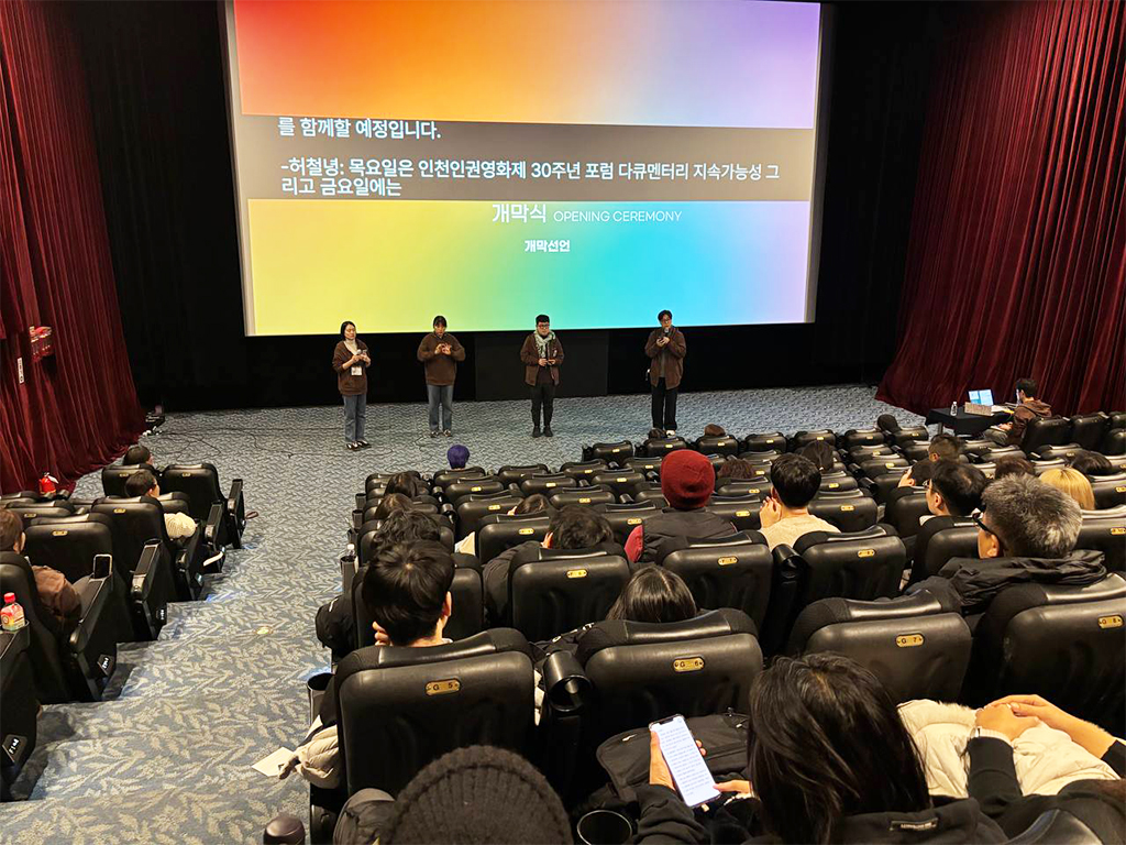 극장 공간 앞 무대 위에 네 명의 사람이 나란히 서서 마이크를 잡고 이야기를 하고 있다. 무대 뒤 스크린에는 무지개색 배경 위 검정색 박스에 실시간 문자통역 글자가 흰색으로 비치고 있다.
: 30회 인천인권영화제 <개막식> 풍경