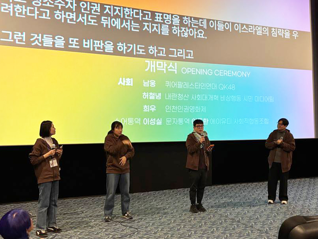 극장 공간 앞 무대 위에 네 명의 사람이 나란히 서서 마이크를 잡고 이야기를 하고 있다. 무대 뒤 스크린에는 무지개색 배경 위 검정색 박스에 실시간 문자통역 글자가 흰색으로 비치고 있다.
: 30회 인천인권영화제 <개막식> 풍경
