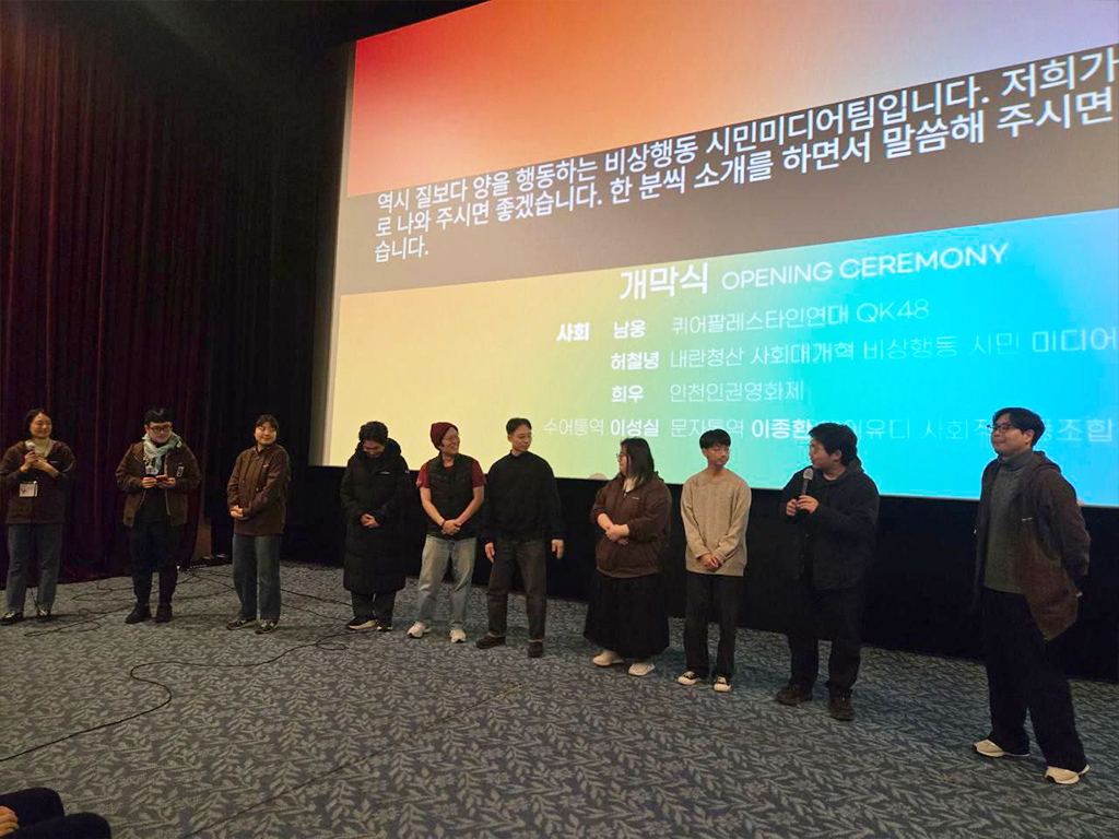 극장 공간 앞 무대 위에 <비상행동 시민미디어팀>열명의 사람이 나란히 서서 마이크를 잡고 이야기를 하고 있다. 무대 뒤 스크린에는 무지개색 배경 위 검정색 박스에 실시간 문자통역 글자가 흰색으로 비치고 있다.
: 30회 인천인권영화제 <개막식> 풍경