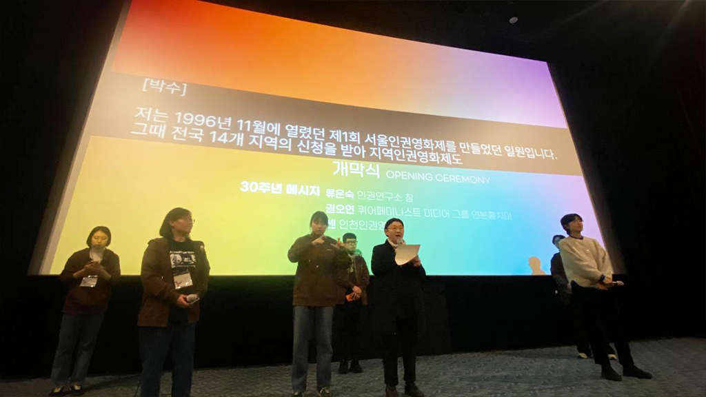 극장 공간 앞 무대 위에 네명의 사람이 나란히 서서 마이크를 잡고 30주년 메세지를 전하고 있다. 무대 뒤 스크린에는 무지개색 배경 위 검정색 박스에 실시간 문자통역 글자가 흰색으로 비치고 있다.
: 30회 인천인권영화제 <개막식> 풍경