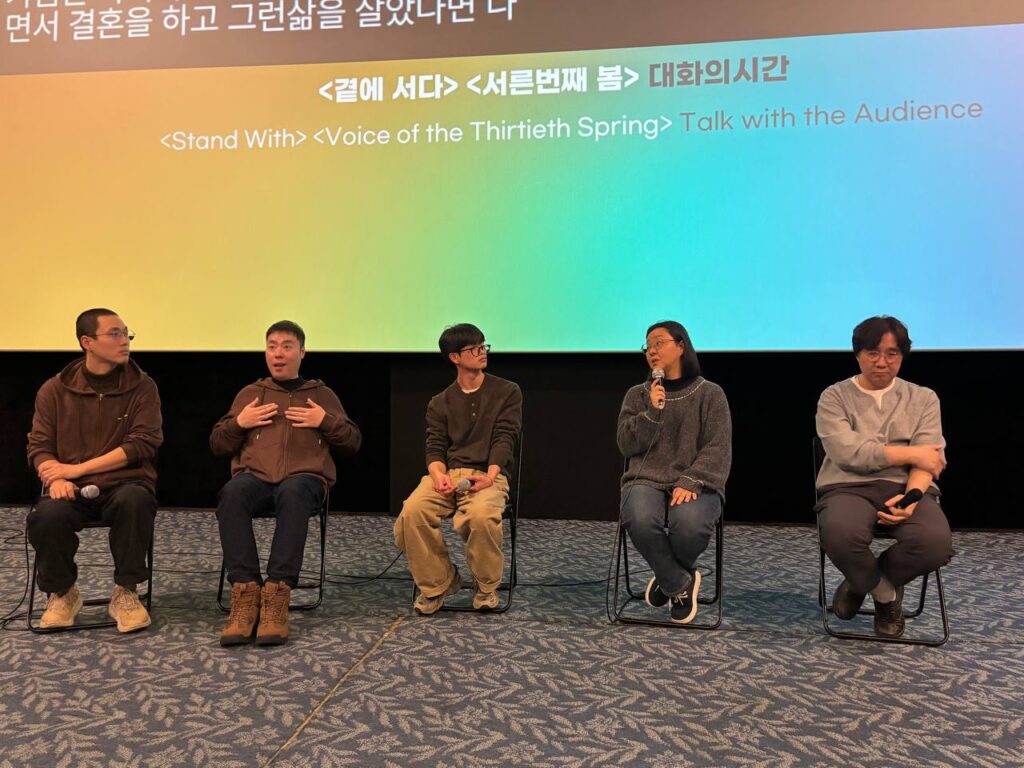 극장 공간 앞 무대 위에 다섯 명의 사람이 앉아 마이크를 잡고 이야기를 하고 있다. 무대 뒤 스크린에는 무지개색 배경 위 검정색 박스에 실시간 문자통역 글자가 흰색으로 비치고 있다.
: 30회 인천인권영화제 <곁에 서다>,<서른 번째의 봄>대화의 시간 풍경