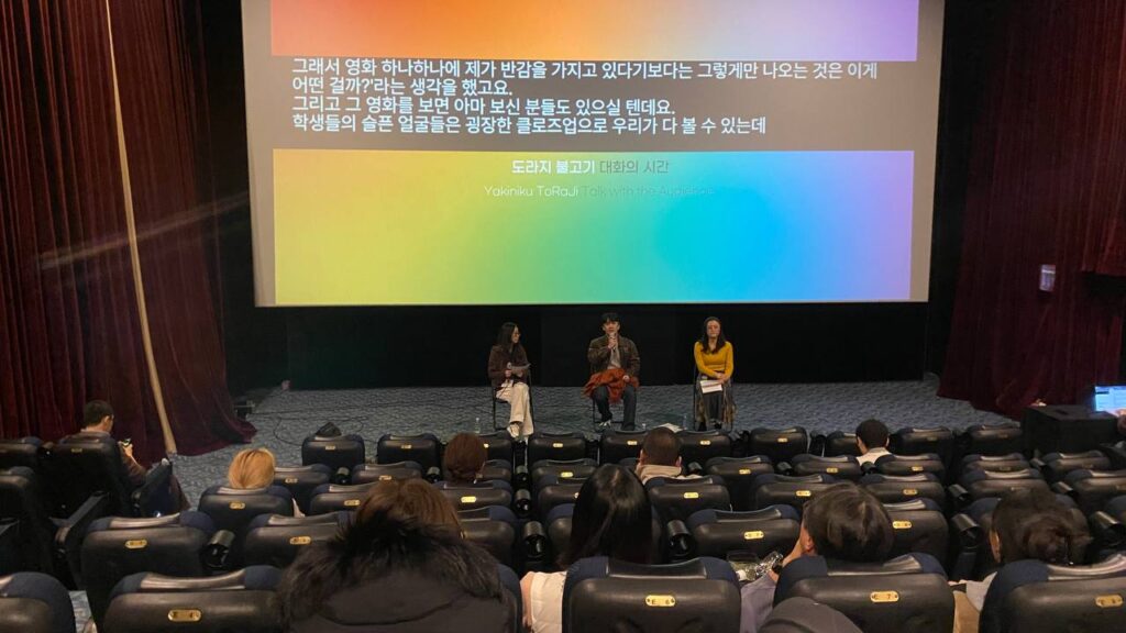 극장 공간 앞 무대 위에 세 명의 사람이 앉아 마이크를 잡고 이야기를 하고 있다. 무대 뒤 스크린에는 무지개색 배경 위 검정색 박스에 실시간 문자통역 글자가 흰색으로 비치고 있다.
: 30회 인천인권영화제 <도라지 불고기>대화의 시간 풍경