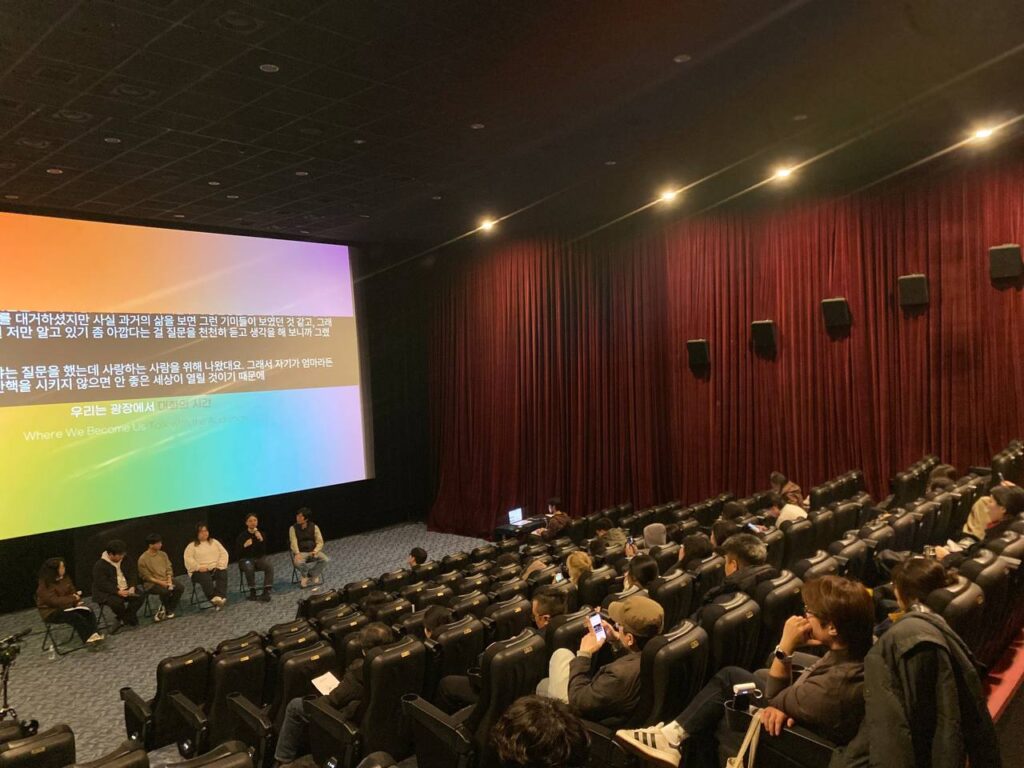 극장 공간 앞 무대 위에 여섯 명의 사람이 앉아 마이크를 잡고 이야기를 하고 있다. 무대 뒤 스크린에는 무지개색 배경 위 검정색 박스에 실시간 문자통역 글자가 흰색으로 비치고 있다.
: 30회 인천인권영화제 <우리는 광장에서>대화의 시간 풍경